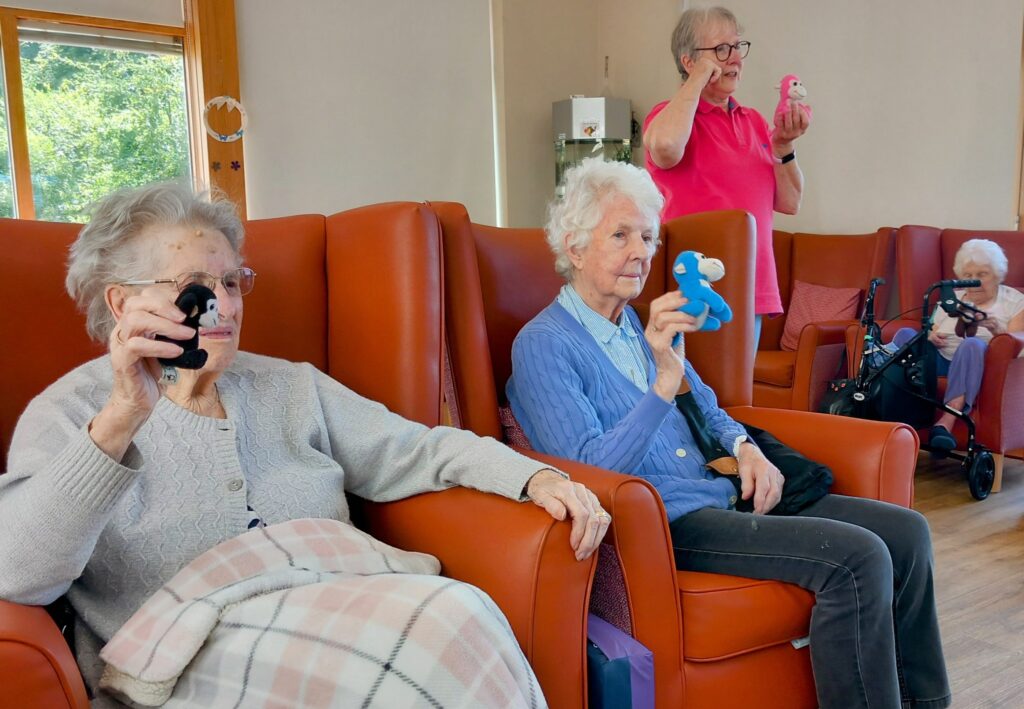 Four elderly women sit in a common room, each holding up a small soft toy, bringing generations together through shared moments and simple pleasures. One woman uses a Zimmer frame in the background.