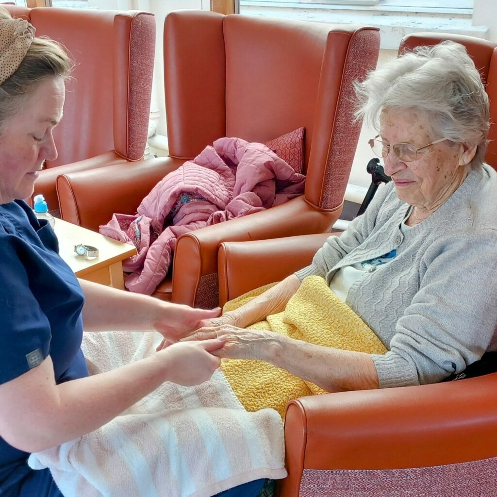 A carer in blue scrubs gently massages the hands of an elderly woman sitting in a chair with a yellow blanket over her lap, providing a relaxing pampering session.