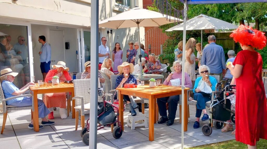 A group of elderly people are seated at outdoor tables, some with walking frames, enjoying a garden party while others stand in the background; someone in a red outfit is addressing the group.