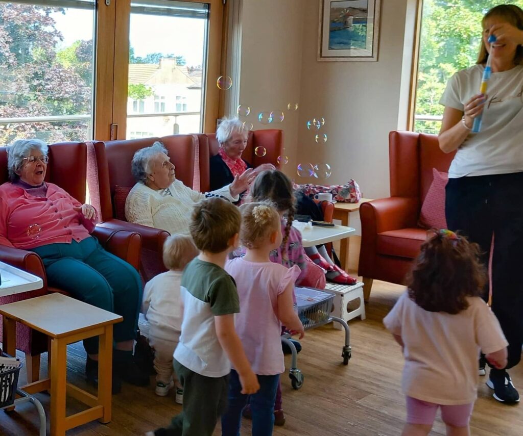 A group of elderly women sit on armchairs while several young children stand nearby; a woman blows bubbles in the brightly lit room at Carries Nursery.