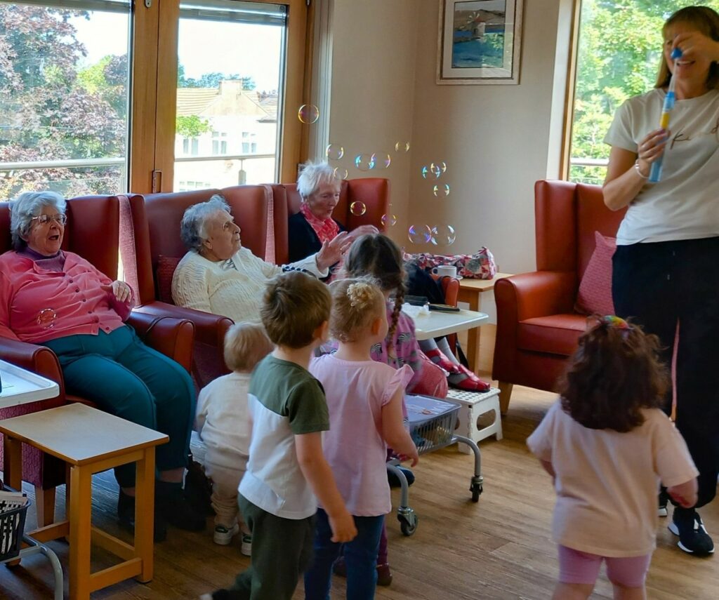 A group of elderly women sit on chairs, while several young children stand and watch a woman blowing bubbles in a brightly lit room.