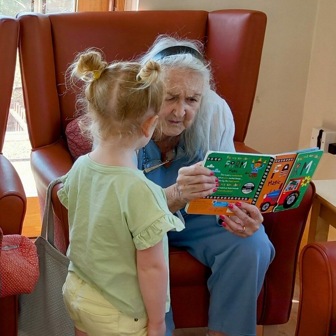 Two elderly women sit in armchairs reading books to a young boy and girl standing in front of them, bringing generations together in a bright room. British English version: Two elderly women sit in armchairs reading books to a young boy and girl who are standing before them, bringing generations together in a bright room.