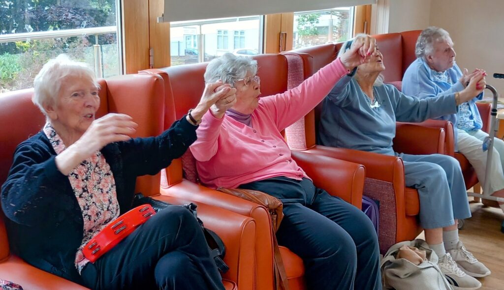 Four elderly women seated in armchairs take part in an activity, raising their arms and holding small objects, in a bright communal room.