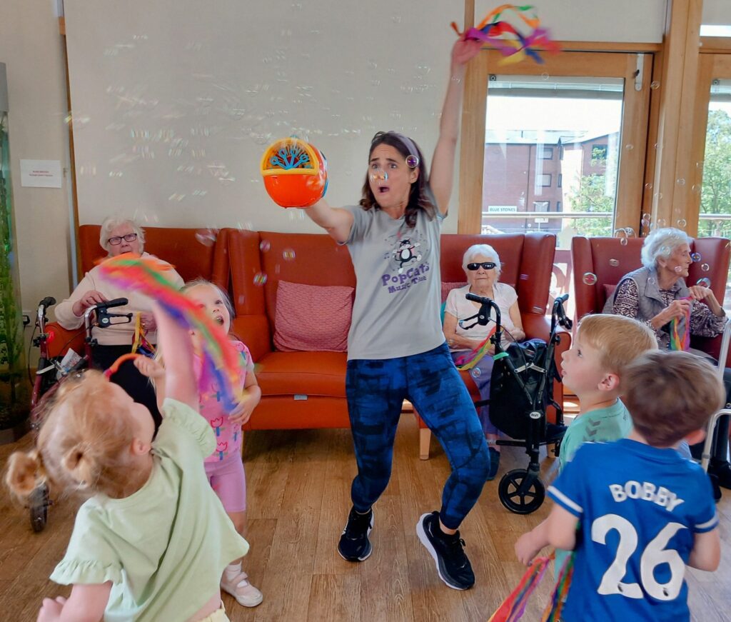 A woman operates a bubble machine as children and elderly adults watch and play with colourful ribbons, bringing generations together in a brightly lit room.