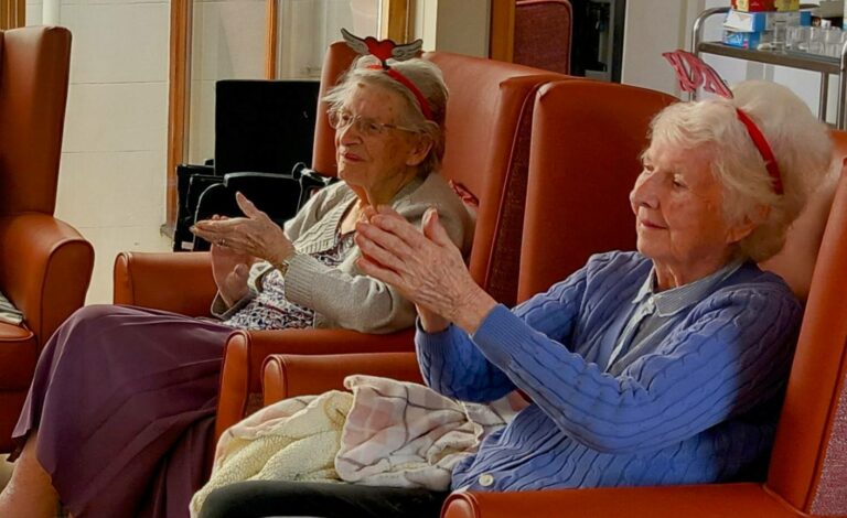 Two elderly women sit in armchairs, wearing red and white headbands, clapping their hands as they share laughter and appear engaged and contented.