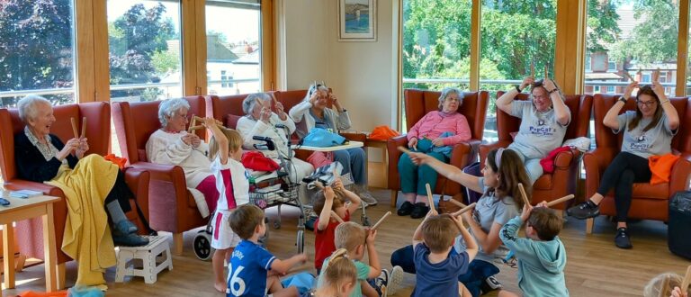 Children and elderly people sit in a circle, following guided movements with their hands, enjoying intergenerational activities in a bright room with large windows and orange chairs.