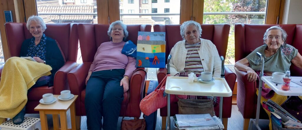 Four elderly women sit in armchairs with drinks and trays at Abbeyfield Lear House; a colourful drawing—perhaps a seaside masterpiece—is displayed between the two women in the centre.