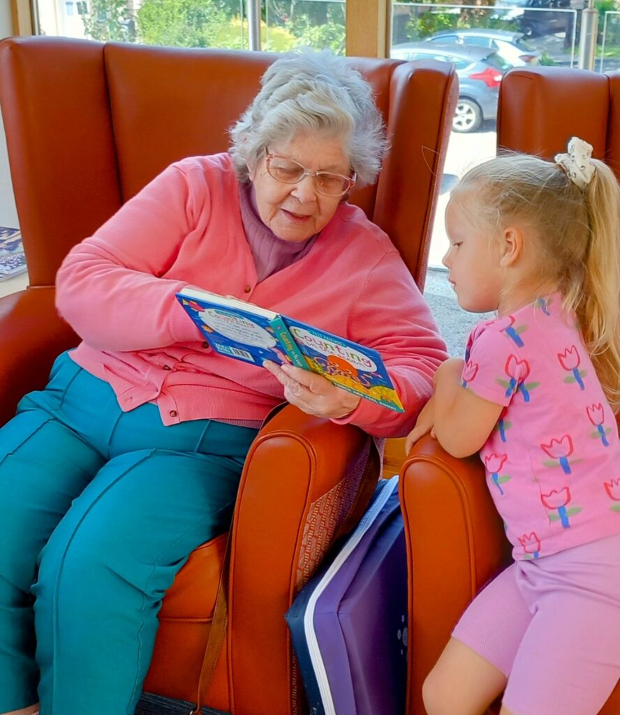An elderly woman reads a childrens book to a young girl sitting beside her in a brightly lit room with large armchairs, beautifully bringing generations together.