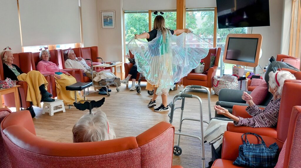 A group of elderly people sit in a lounge, sharing laughter as they watch a performer with shiny wings dance in the centre of the room.