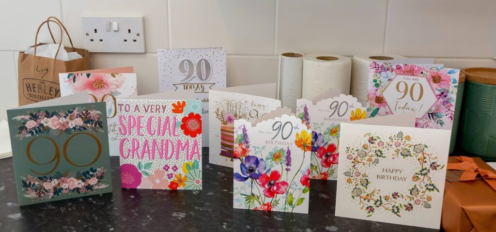 A row of birthday cards, many featuring the number 90 and floral designs, are displayed on a kitchen worktop, setting the scene for a special day of celebration. Kitchen roll and a paper bag sit in the background.