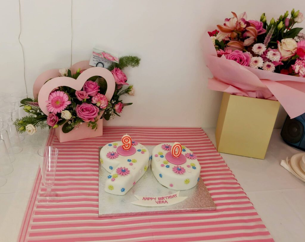 Two heart-shaped cakes with “90” candles, a bouquet of pink flowers, and a heart-shaped floral arrangement set the scene for Vera’s special day on a pink-striped tablecloth—perfect for a garden party.