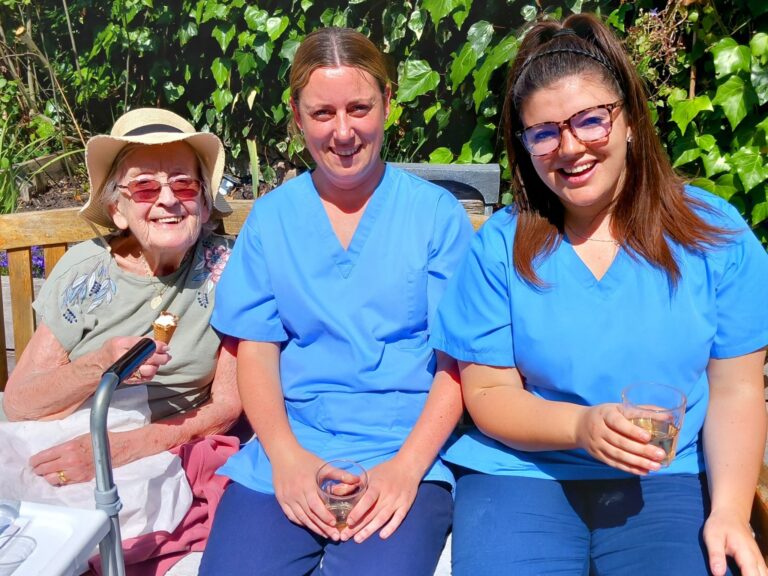 An elderly woman with a walking frame enjoys ice cream outdoors beside two women in blue scrubs, each holding a drink, during Vera’s special day, with lush greenery in the background. Rewritten in British English: An elderly woman with a walking frame enjoys ice cream outdoors next to two women in blue scrubs, each holding a drink, during Vera’s special day, with lush greenery in the background.