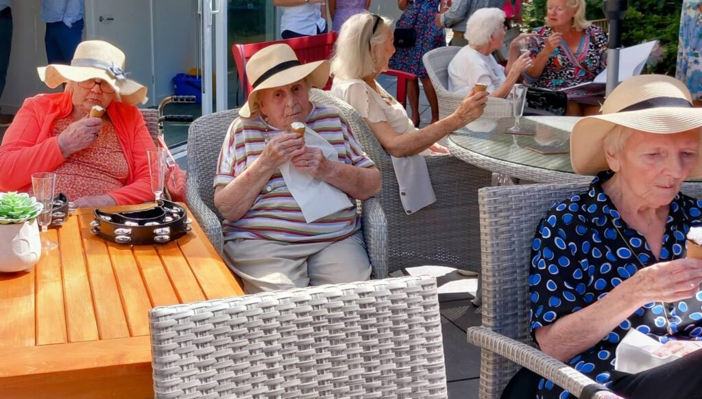 Elderly women wearing hats are seated outside at tables, enjoying ice cream cones on a sunny day. It appears to be a garden party, with people in the background also sitting and socialising.