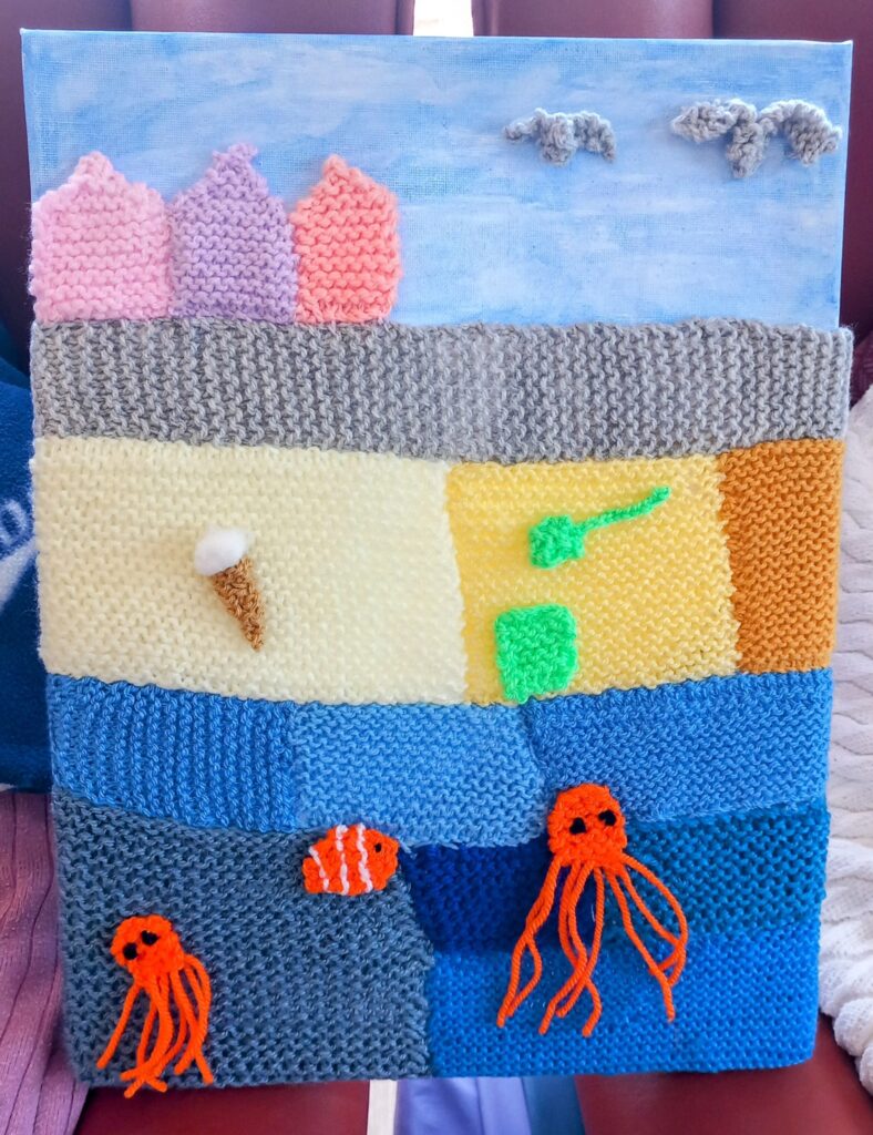 A seaside masterpiece in knitting, this artwork features three pink houses, clouds, an ice cream cone, a green spade, and two orange jellyfish under the water.