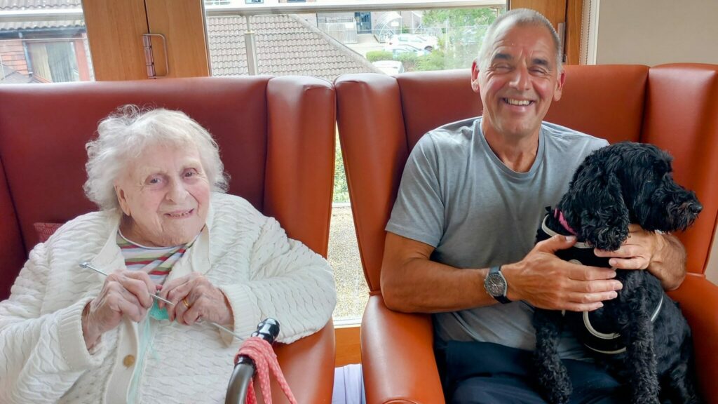 An elderly woman sits smiling, knitting beside a man holding a black dog; both are seated in orange armchairs by a window at Abbeyfield Lear House.