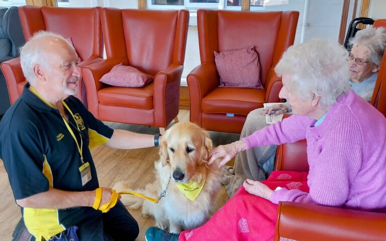 An elderly woman strokes a golden retriever therapy dog while a man holds its lead in a common room, with another elderly woman seated nearby.
