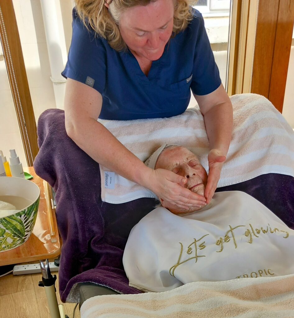 A woman in a blue top gives an elderly person a relaxing facial massage during a pamper session as they recline with a towel and a “Let’s get glowing” bib. A bowl and skincare products are visible nearby.