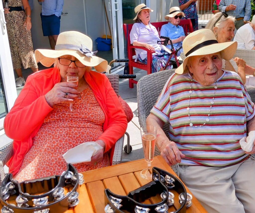 Two elderly women in sun hats enjoy tambourines and sparkling drinks at a lively garden party; other pensioners relax in the background, celebrating Veras Special Day.