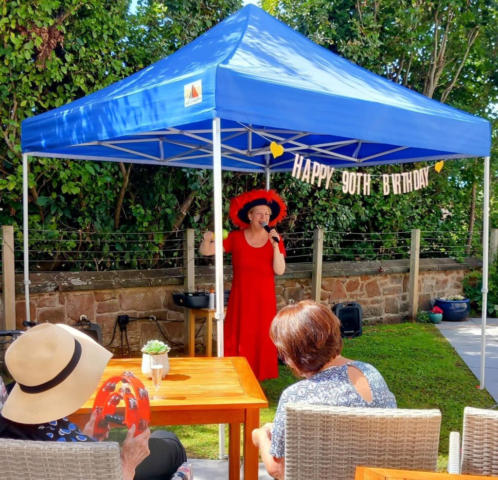 Vera, dressed in a red dress and hat, sings beneath a blue canopy with a ‘Happy 90th Birthday’ banner, while guests enjoy her special day at a charming outdoor garden party.