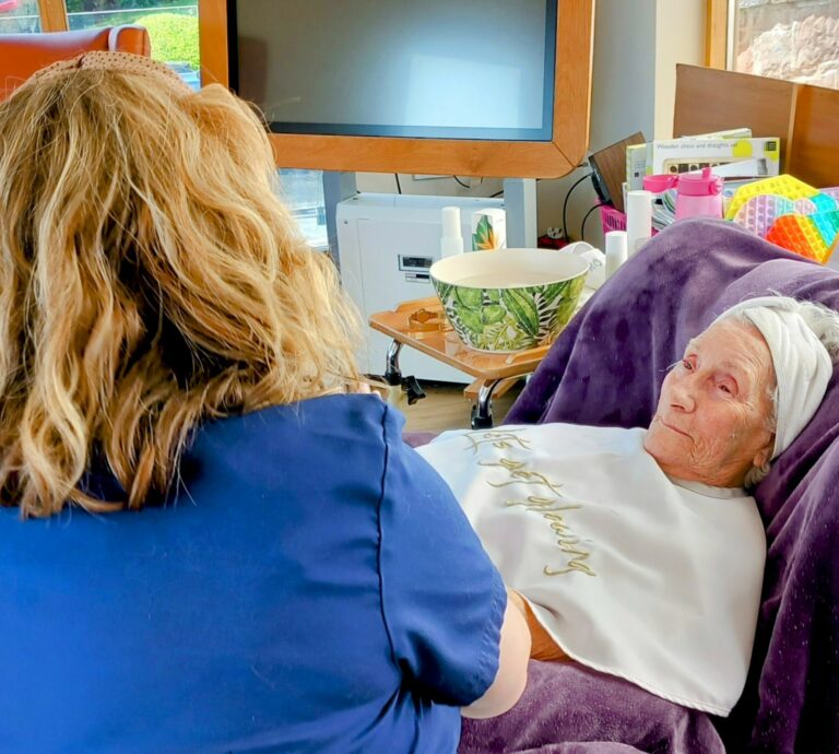 An elderly woman reclines on a chair with a towel wrapped around her head, enjoying a pampering session as another person, seen from behind, attends to her.