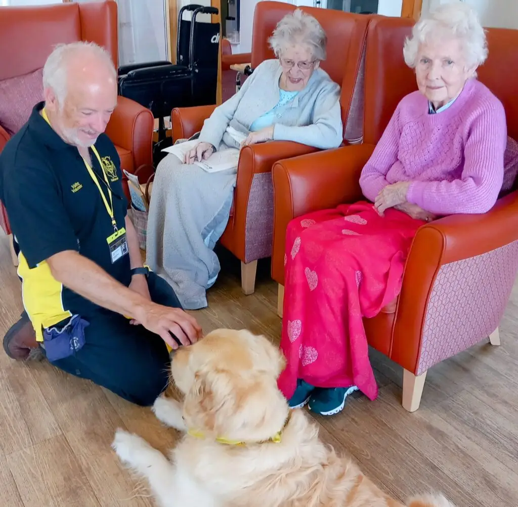 A man sits on the floor stroking a golden retriever while two elderly women sit in armchairs, one wearing a pink jumper and red blanket, the other holding papers.