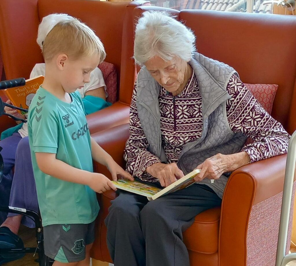 A young boy stands next to an elderly woman seated in an armchair as they look together at a book she is holding open on her lap, sharing a special moment between generations.