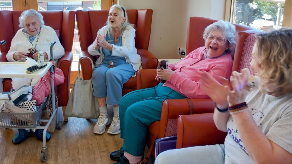 Four elderly women sit on chairs indoors, smiling and clapping together as bright daylight streams through the windows—celebrating generational connection in a warm, joyful atmosphere.