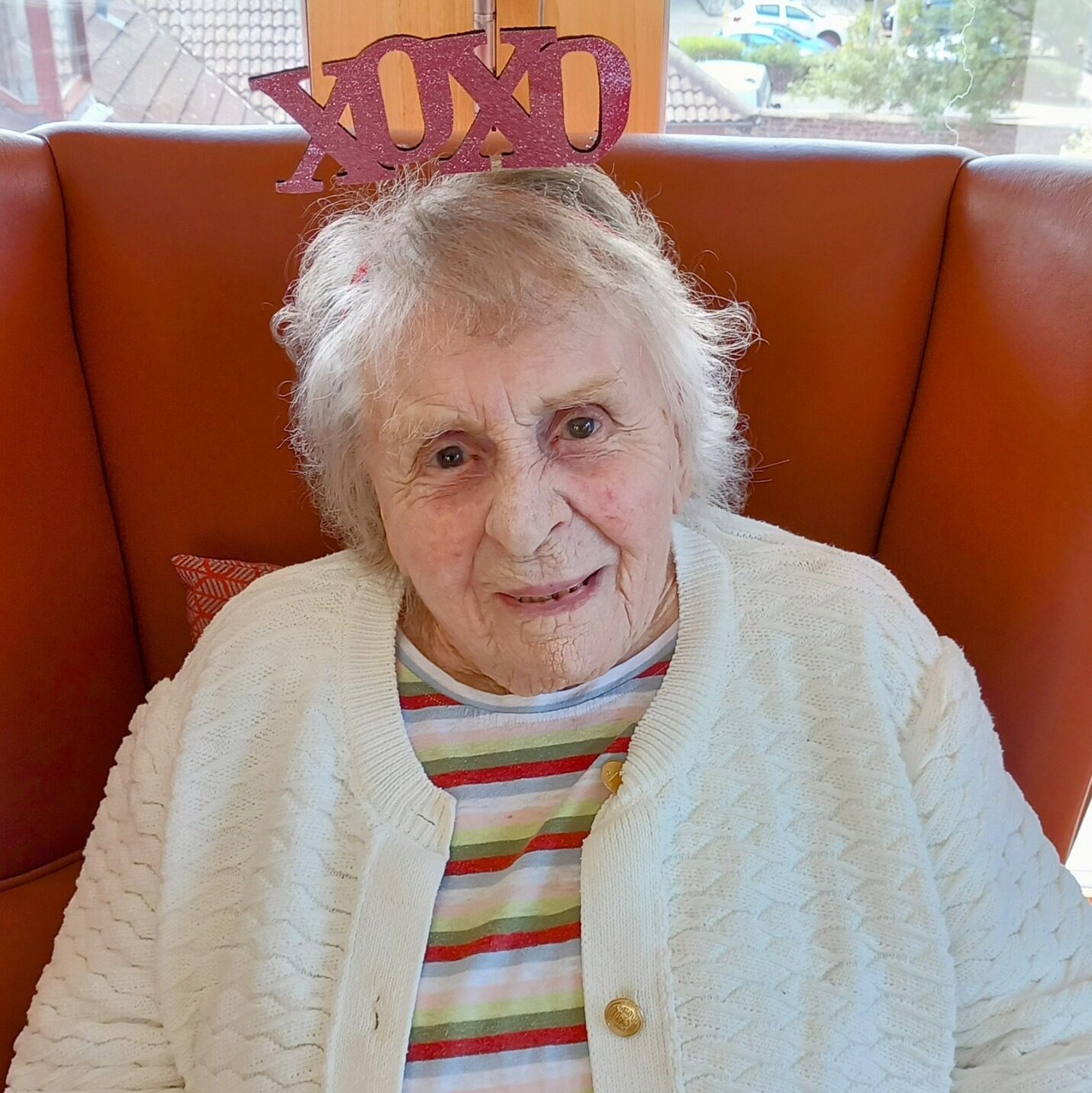 Elderly woman with white hair sits in an orange armchair, wearing a striped top, a white cardigan, and a headband that reads XOXO, sharing love and warmth with her bright smile.