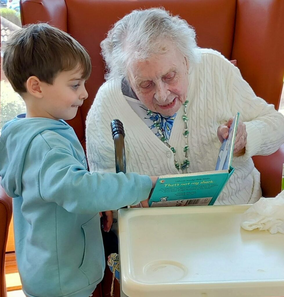 An elderly woman and a young boy sit together; she reads from a children’s book while he points to something on the page, highlighting the warmth of generational bonding.