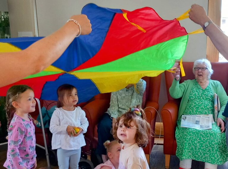 Several children and elderly adults are playing together with a colourful parachute at Carries Nursery; one older woman sits smiling and holding a newspaper.