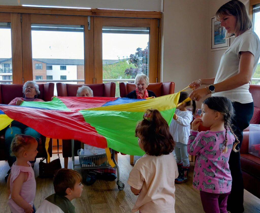 Children and elderly people hold a colourful parachute together indoors at Caroline’s Childminding, enjoying a lively group activity.