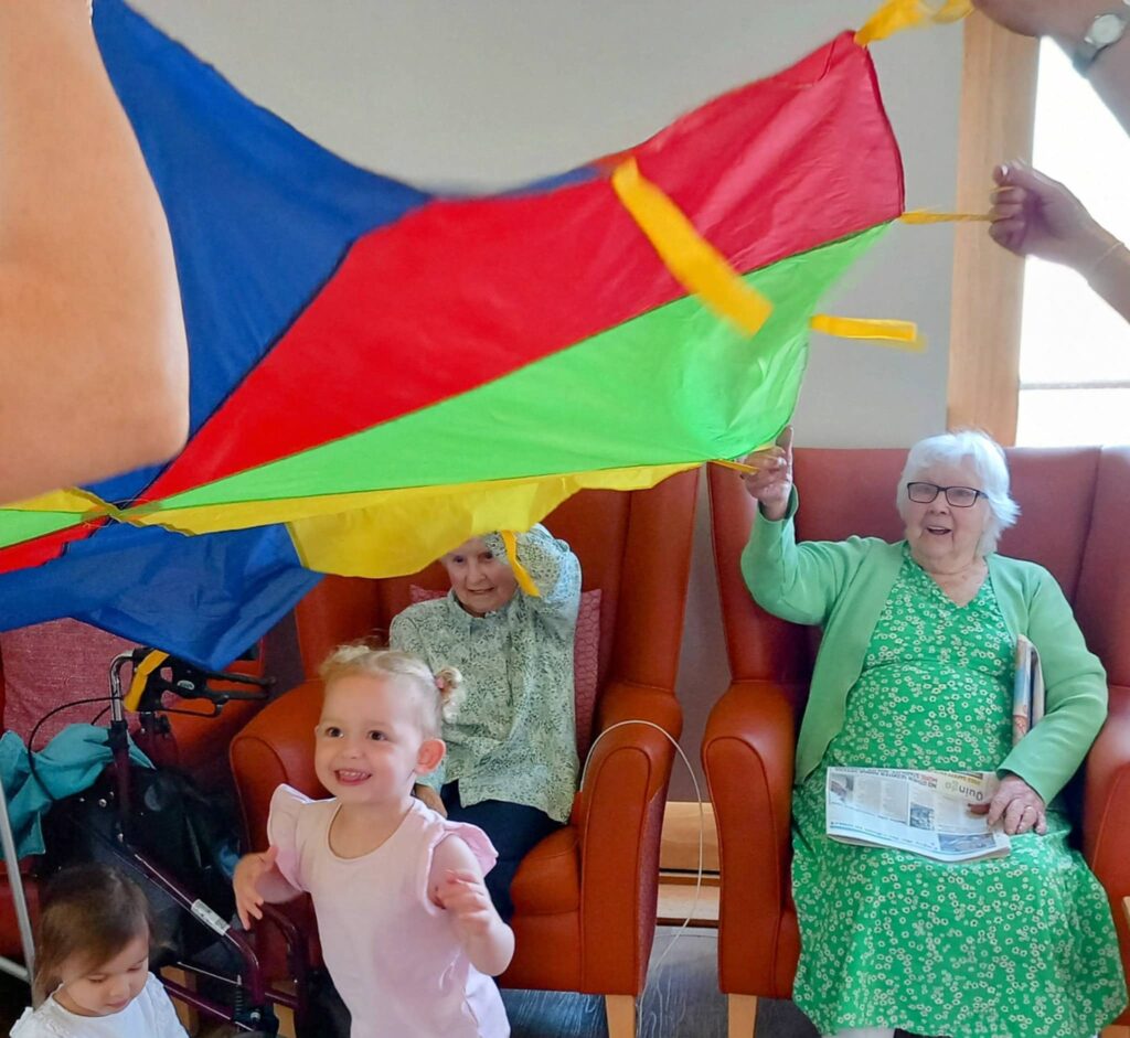 Two elderly women and two young children play with a colourful parachute indoors at Caroline’s Childminding; one elderly woman holds a newspaper and smiles.