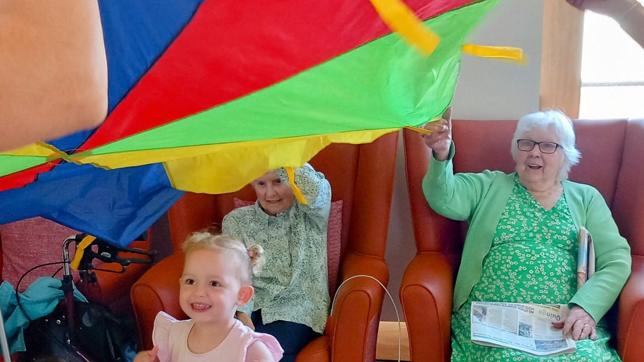 Young children and elderly women sit together indoors, playing with a colourful parachute. One woman holds a newspaper; another smiles, wearing a green patterned jumper.