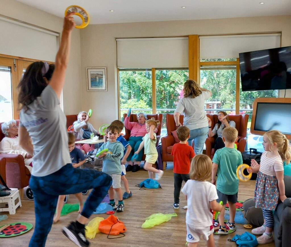Adults and children take part in a lively indoor group activity, with some playing musical instruments, while seated adults observe and interact—bringing generations together in a vibrant, intergenerational setting.