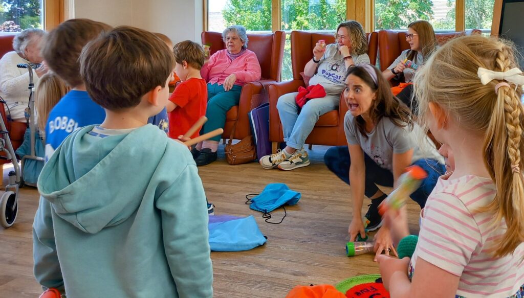Bringing Generations Together, children play musical instruments in a brightly lit room while adults watch from armchairs in the background.