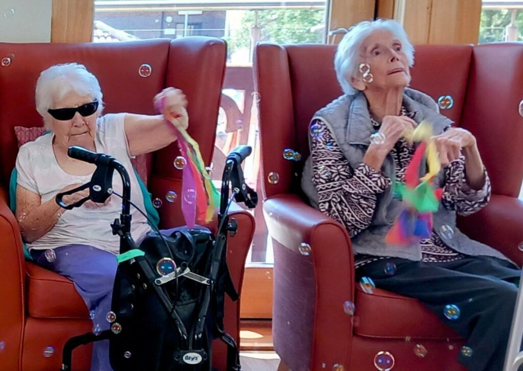 Two elderly women sit in armchairs, holding colourful ribbon wands and surrounded by bubbles. One wears sunglasses and has a walking frame in front of her. Both enjoy the moment, highlighting the joy of generational bonding.