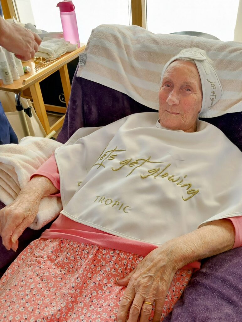 Elderly woman enjoying a pampering session, seated in a chair with a towel on her head and a bib that reads lets get glowing, wearing a pink top and patterned trousers.