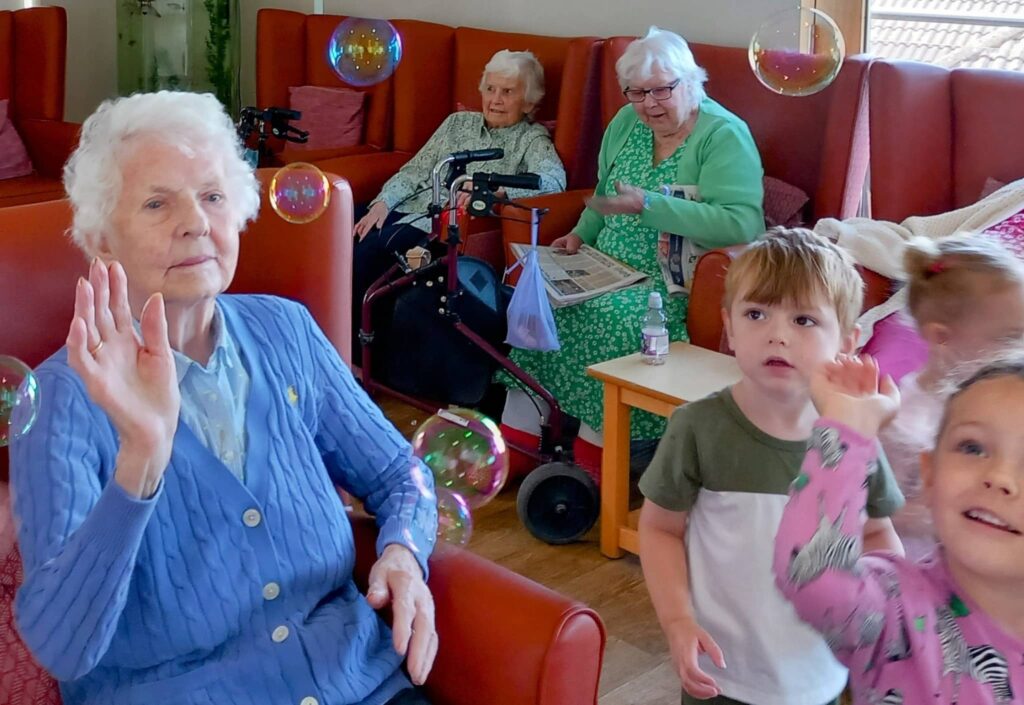 Elderly women sit in a lounge area while young children from Caroline’s Childminding play with soap bubbles nearby.