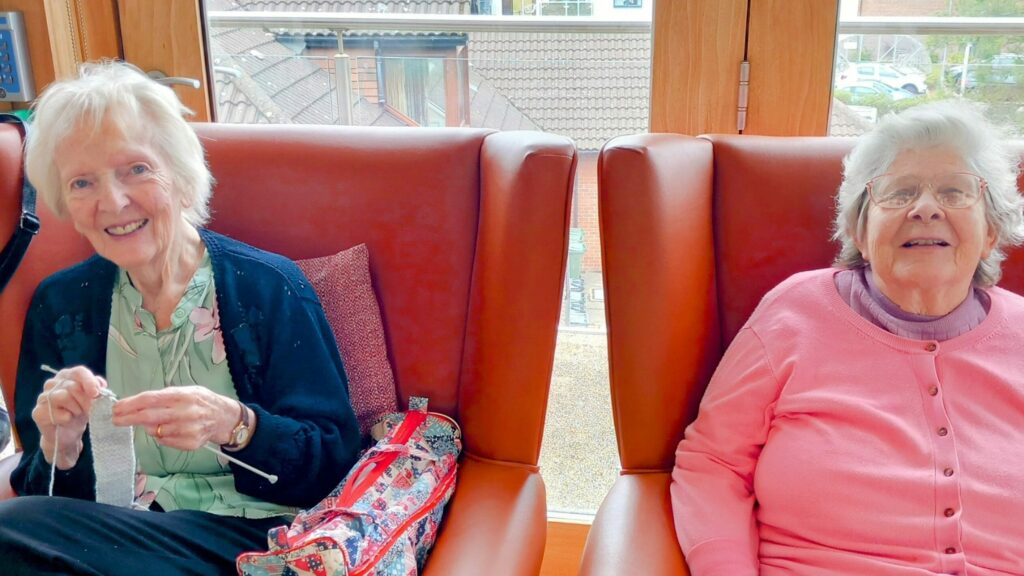Two elderly women are seated in armchairs at Abbeyfield Lear House, one knitting and smiling, the other wearing glasses and a pink cardigan; both are looking at the camera.