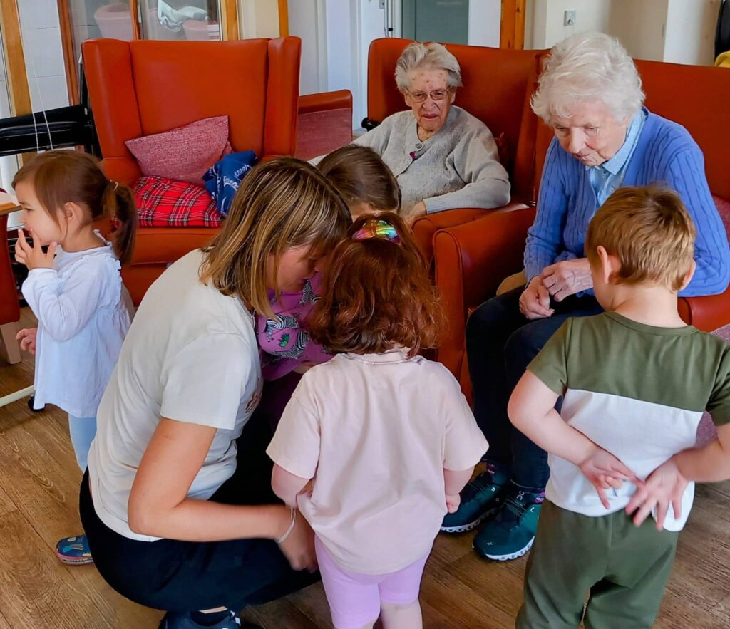 Several young children and a woman gather in a group while two elderly women sit in armchairs observing them—a warm, cosy scene at Caroline’s Childminding.