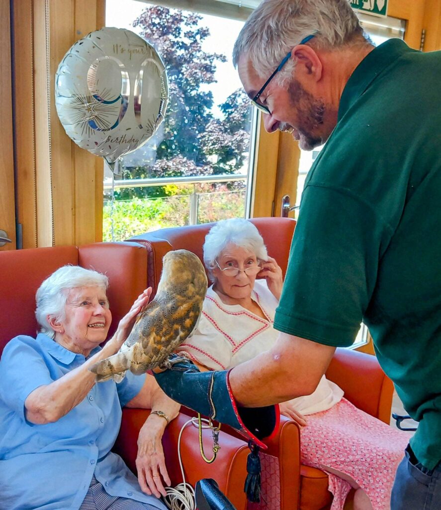 A man holding a barn owl allows two elderly women on a sofa to interact with their feathered companion during an afternoon event, with a 90th birthday balloon glowing in the bright room.