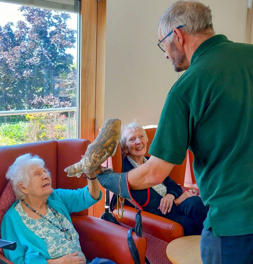 An elderly woman in a care home sits in a chair and strokes a large tortoise held by a man wearing gloves during an animal event; another woman sits nearby, smiling.
