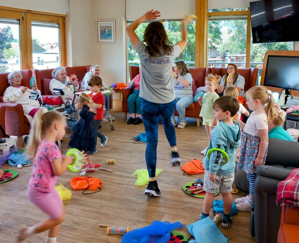 A group of children dance and play with colourful toys while older adults watch, bringing generations together in a bright room with large windows and wooden floors.