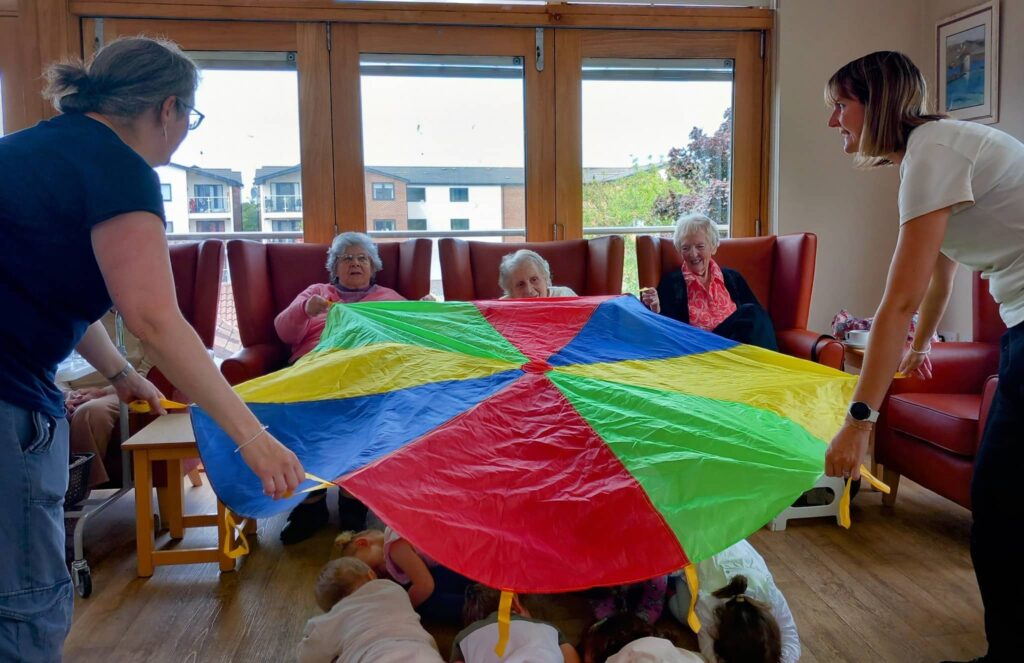 Four elderly women sit on chairs holding a colourful parachute, with two younger adults at either end, while children from Caroline’s Childminding play underneath in a bright community room.