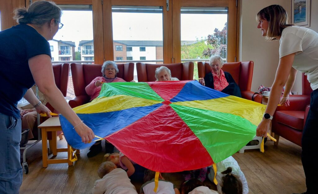 Four elderly women are seated in chairs while two carers and several children hold a colourful parachute indoors, taking part in a group activity.