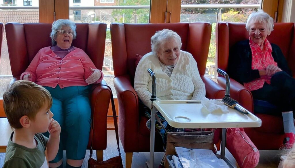 Three elderly women are seated in armchairs, smiling, with a young boy from Caroline’s Childminding in the foreground. A tray with items and walking aids are visible between the women.