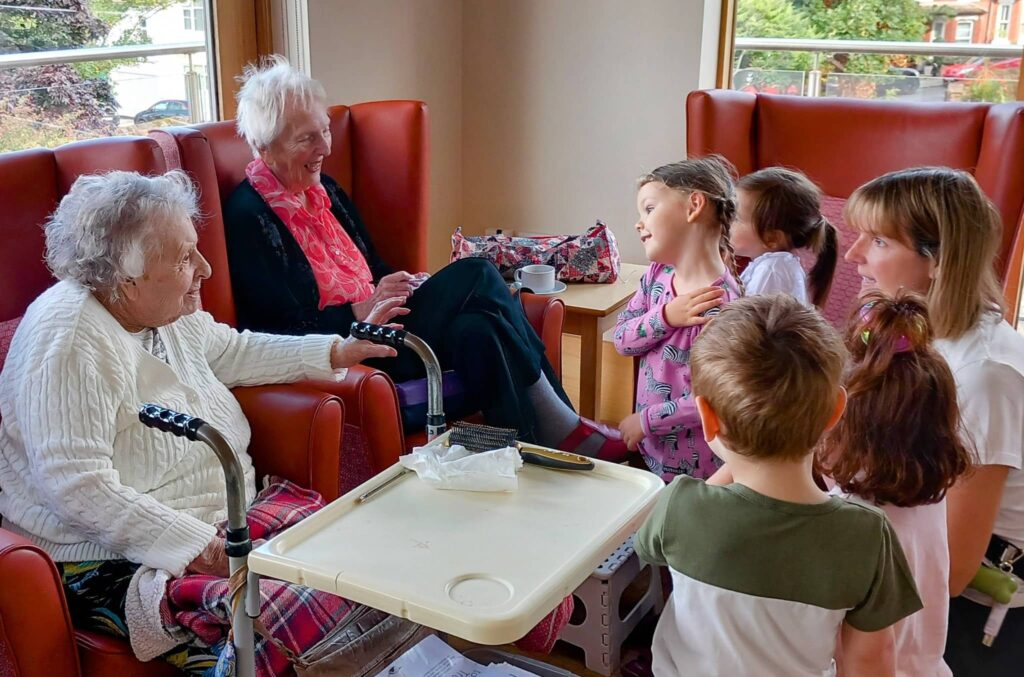 Two elderly women sit in chairs and engage with a group of young children and an adult woman in a brightly lit room with large windows at Caroline’s Childminding.