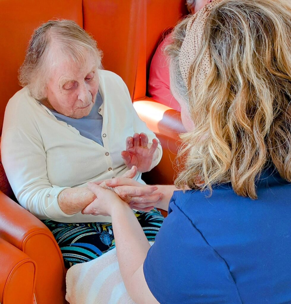 An elderly woman enjoys a pampering session, seated in an orange chair as another woman gently holds and cares for her hands.