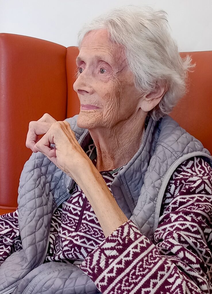 An elderly woman with white hair sits on an orange chair, wearing a patterned jumper and a quilted gilet, looking to the side with a calm expression.