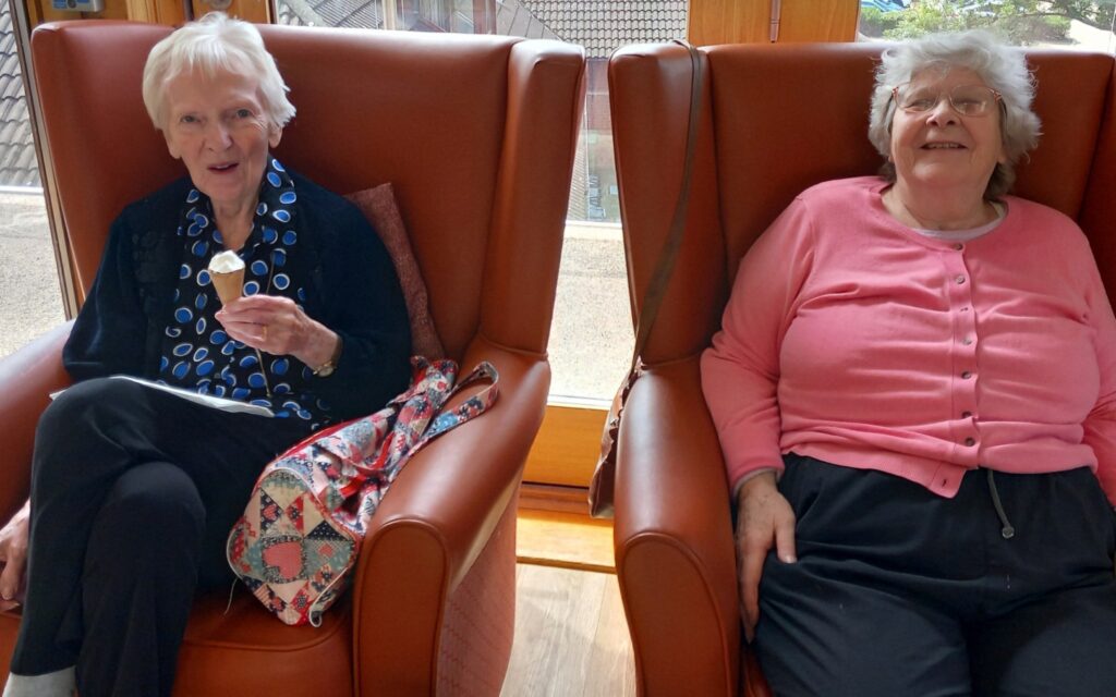 Two elderly women enjoy a pampering session in brown armchairs by a window; one is eating an ice cream cone, while the other relaxes with her hands in her lap.